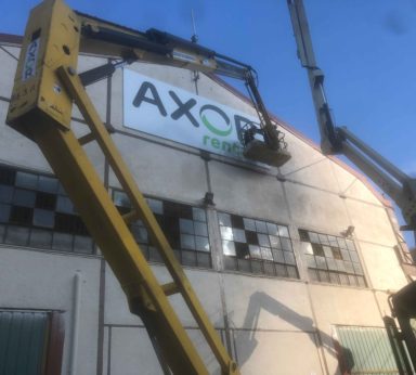 Operarios con grúa colocando cartel de logo de la empresa Axor Rentals en parte superior de pabellón industrial