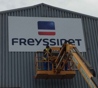 Operarios con grúa colocando cartel de logotipo de la empresa Freyssinet en parte superior de pabellón industrial